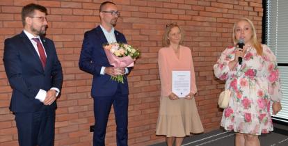 Cztery osoby stoją przed ceglaną ścianą podczas uroczystego podziękowania. Jedna z kobiet trzyma dyplom, mężczyzna bukiet kwiatów, a druga kobieta przemawia do mikrofonu
