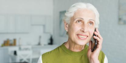 Uśmiechnięta seniorka o siwych włosach rozmawia przez telefon komórkowy. Kobieta przebywa w jasnym, domowym wnętrzu