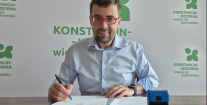 Burmistrz siedzi przy biurku i podpisuje dokumenty, a na stole obok znajdują się pieczątka oraz dokumentacja.