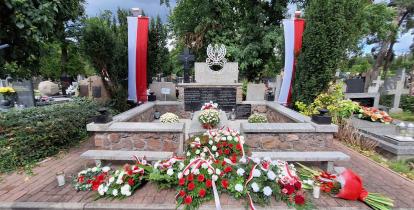 Pomnik wojenny na cmentarzu udekorowany biało-czerwonymi wieńcami i wiązankami kwiatów, fluktuujący dwoma polskimi flagami po bokach. Na płycie widnieje godło Polski oraz napis „Chwała Bohaterom”.