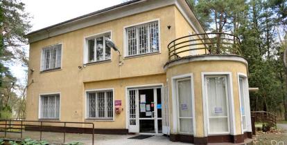 Budynek przychodni wśród drzew, z żółtą elewacją, dużymi oknami z kratami oraz zaokrąglonym wejściem z podjazdem dla osób z niepełnosprawnościami.