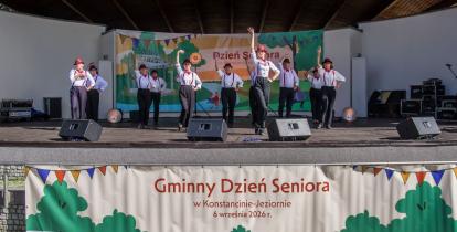 Występ artystyczny seniorów na scenie amfiteatru podczas Gminnego Dnia Seniora w Konstancinie-Jeziornie. Grupa tancerzy w białych koszulach, ciemnych spodniach, czerwonych szelkach i kapeluszach wykonuje układ taneczny z uniesionymi rękami. Z przodu sceny wisi duży baner z napisem „Gminny Dzień Seniora w Konstancinie-Jeziornie”.