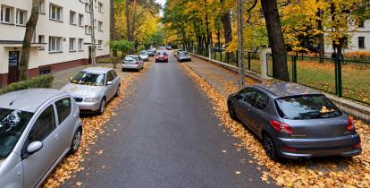 Jesienna, zadrzewiona ulica z zabudową mieszkaniową, chodnikami, latarniami i zaparkowanymi samochodami.