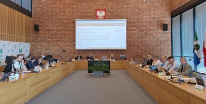 Grupa osób siedzi za stołami na sali posiedzeń
