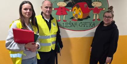 Trzy osoby stoją na korytarzu przy ściennej ilustracji przedstawiającej chatkę w lesie z postaciami z bajki. Dwie osoby mają na sobie żółte kamizelki odblaskowe, jedna trzyma czerwony segregator.