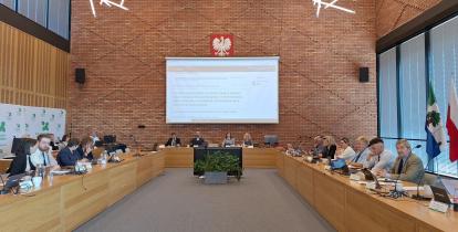 Grupa osób siedzi za stołami na sali posiedzeń