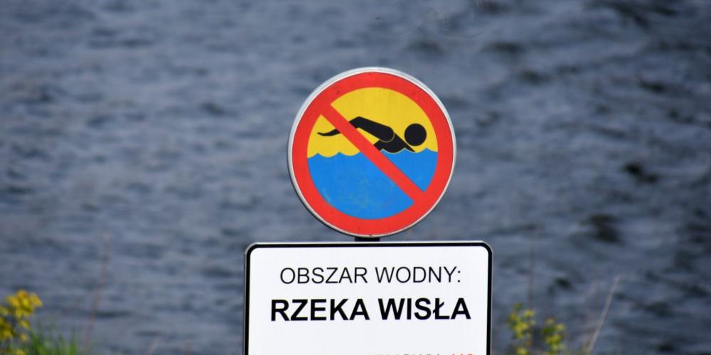 Znak zakazujący kąpieli z tabliczką informacyjną, na której jest tekst: obszar wodny – rzeka Wisła, telefon alarmowy 112. W tle nieostre fale płynącej rzeki.  