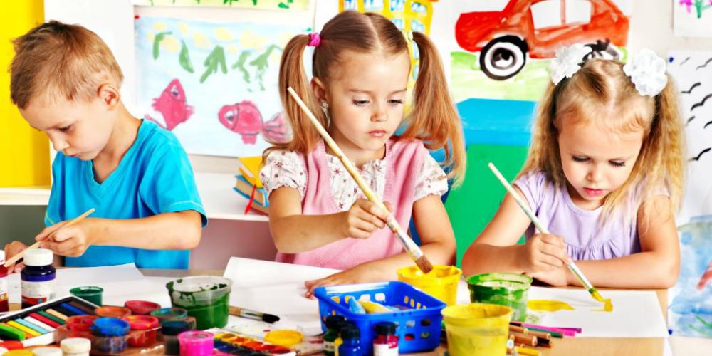 Sala przedszkolna, przy stoliku siedzą dzieci: dwie dziewczynki i jeden chłopiec. Na stole leżą farby do malowania. Wszyscy trzymają pędzle w rękach i coś tworzą. 
