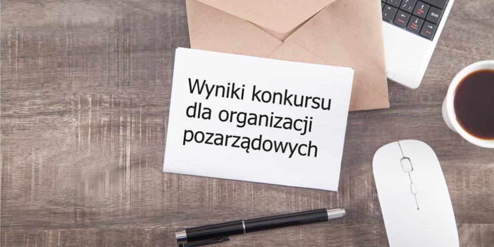 Na biurku stoi komputer, myszka i kubek z kawą na klawiaturze leży otwarta koperta i list