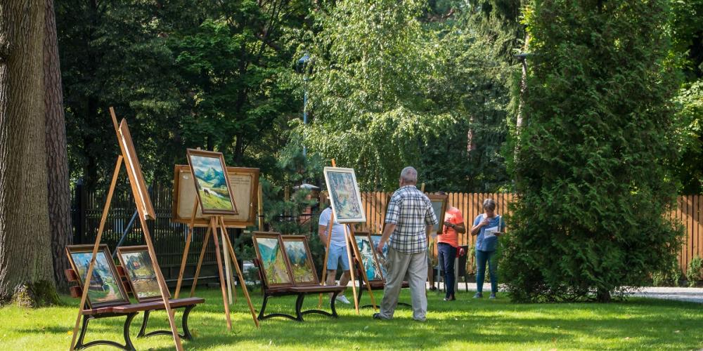 Ogród, na trawie stoją sztalugi drewniane a na nich obrazy. W tle widać ludzi i wysokie drzewa. 