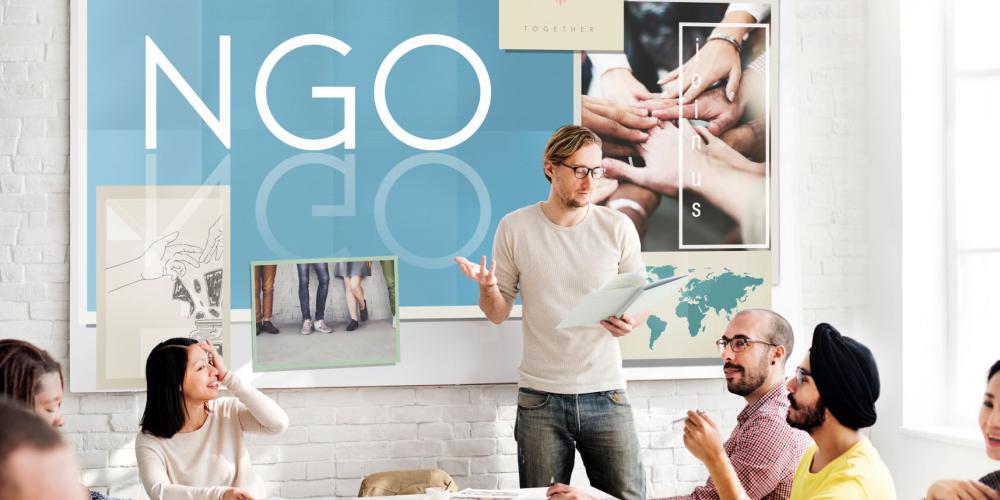 Kilka osób siedzi przy stole i dyskutują. W tle na ścianie widać napis NGO.