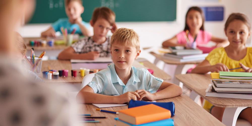 W klasie nauczycielka stoi przy tablicy, dzieci siedzą w ławkach i zgłaszają się do odpowiedzi