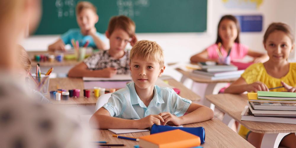 W klasie nauczycielka stoi przy tablicy, dzieci siedzą w ławkach i zgłaszają się do odpowiedzi