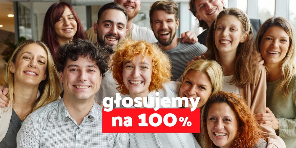 Grupa uśmiechniętych ludzi pozuje razem do zdjęcia, a na pierwszym planie widnieje hasło „głosujemy na 100%” na czerwono-białym tle.