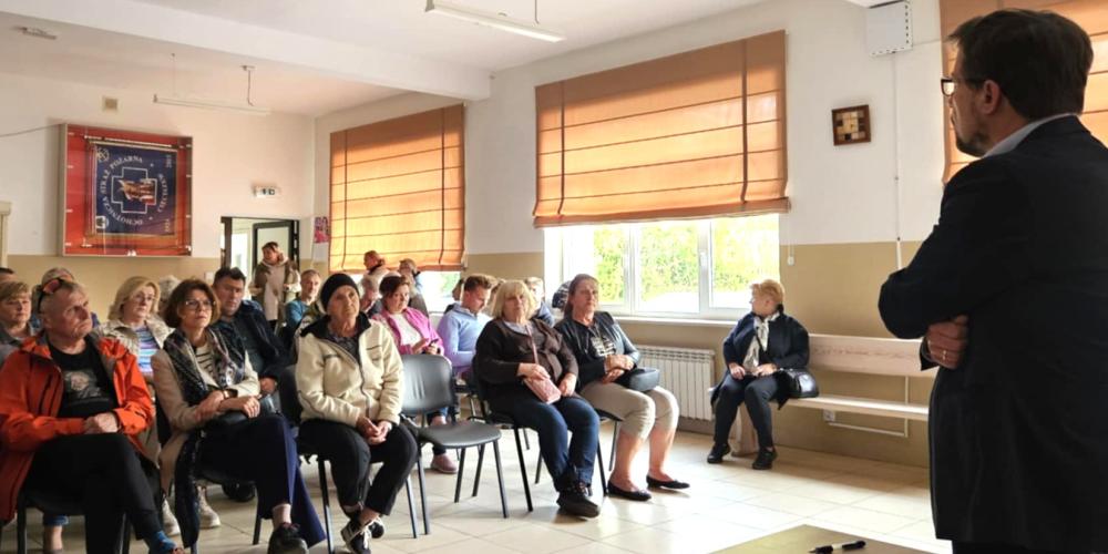Mężczyzna w marynarce przemawia do zgromadzonych mieszkańców siedzących przy stołach, podczas spotkania w świetlicy wiejskiej.