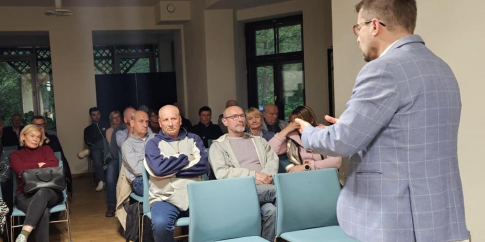 Mężczyzna w marynarce przemawia do zgromadzonych mieszkańców siedzących przy stołach, podczas spotkania w świetlicy wiejskiej.