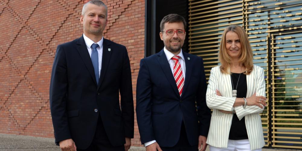 Na zdjęciu trzech uśmiechniętych przedstawicieli samorządu gminy Konstancin-Jeziorna pozuje przed nowoczesnym budynkiem z ceglaną i przeszkloną elewacją.