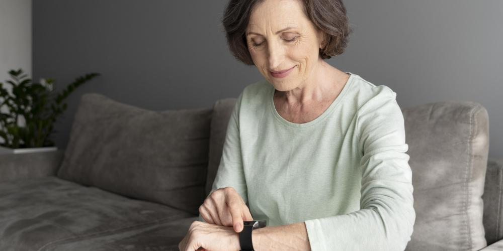 Starsza kobieta siedzi na sofie i uśmiechając się, obsługuje opaskę sportową lub smartwatch na swoim nadgarstku.