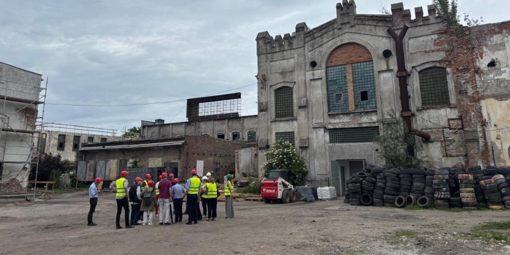 Grupa osób w kamizelkach odblaskowych przed budynkiem pofabrycznym.