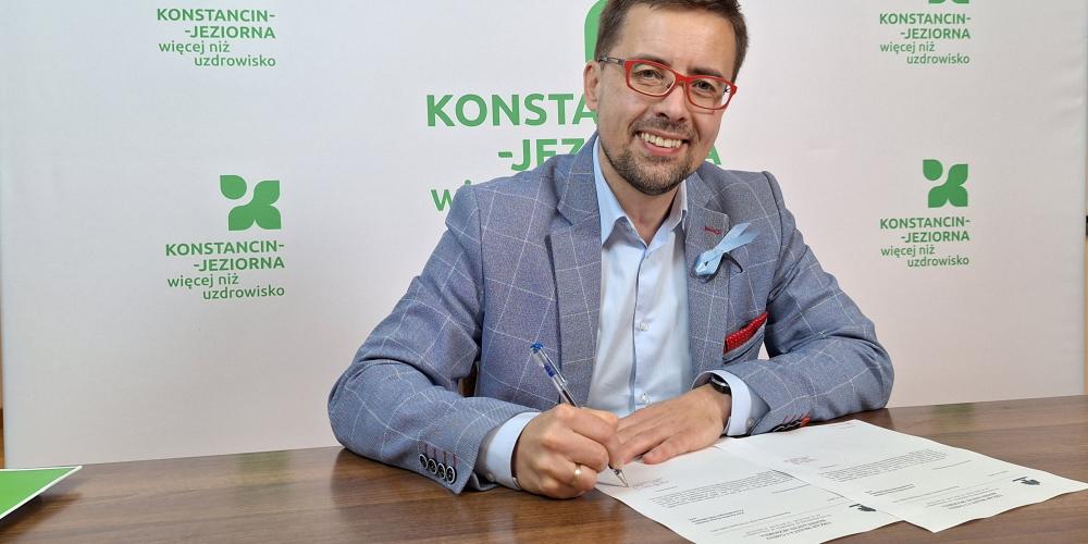 Uśmiechnięty mężczyzna w okularach i eleganckiej marynarce siedzi przy biurku i podpisuje dokumenty na tle banneru promocyjnego miasta.