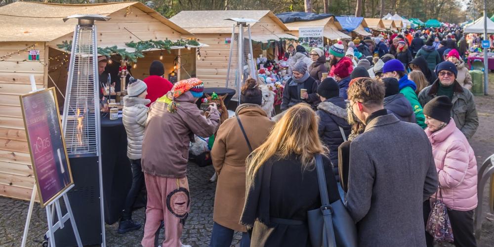 Ludzie spacerują między drewnianymi stoiskami z ozdobami i świątecznymi produktami podczas jarmarku w parku otoczonym drzewami.