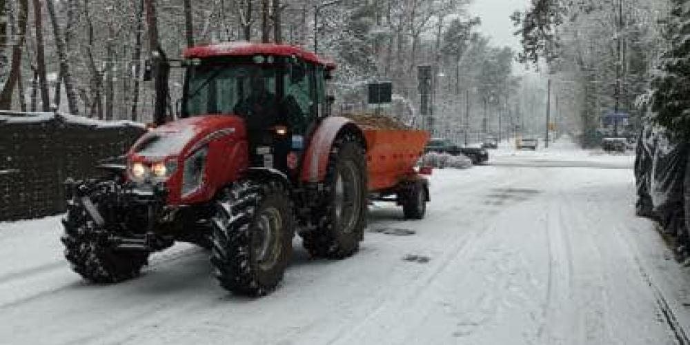 Na ośnieżonej ulicy jedzie czerwony traktor z posypywarką, wykonując zimowe prace utrzymaniowe w otoczeniu drzew i zaparkowanych samochodów.