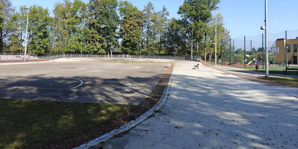 Szkolne boisko z asfaltową nawierzchnią i bramkami, położone obok budynku szkoły, otoczone zielenią i oświetleniem.