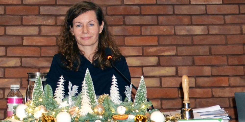 Uśmiechnięta kobieta za stołem prezydialnym, na tle ściany z czerwonej cegły.