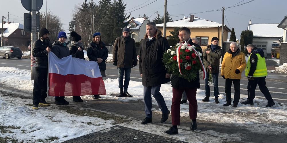 Grupa osób podczas zimowej uroczystości składa kwiaty pod pomnikiem, jedna z nich niesie wieniec, a w tle widoczna jest flaga Polski oraz zabudowa jednorodzinna przy drodze.