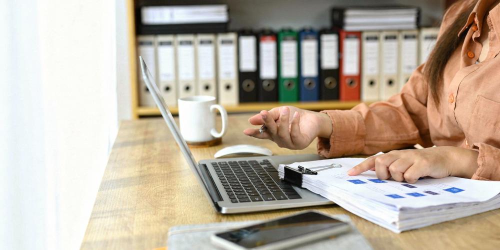 Kobieta siedzi przy biurku i analizuje dokumenty finansowe, pracując na laptopie w biurze z segregatorami na półkach w tle.