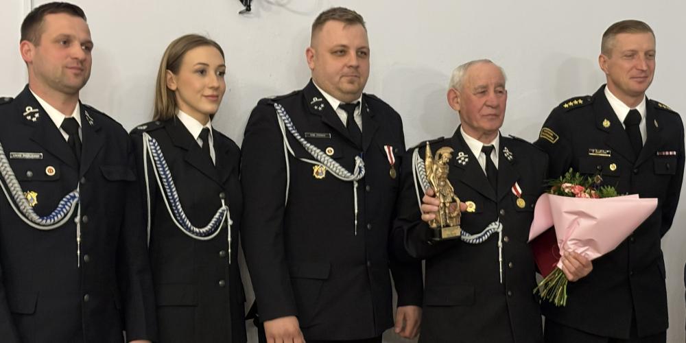 Grupa strażaków w galowych mundurach stoi obok siebie podczas uroczystości, a jeden z nich trzyma statuetkę, inny bukiet kwiatów.