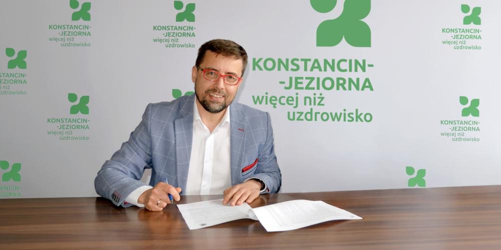 Mężczyzna w okularach podpisuje dokumenty przy stole na tle ścianki z logo „Konstancin-Jeziorna – więcej niż uzdrowisko”.