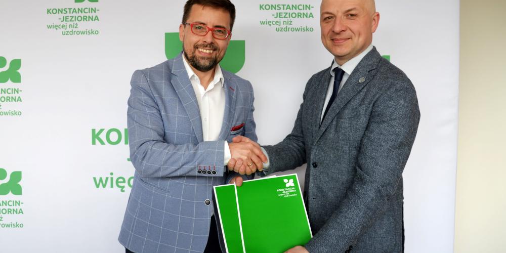 Dwóch mężczyzn w garniturach uściskiem dłoni potwierdza podpisanie dokumentu na tle ścianki z logo Konstancina-Jeziorny.
