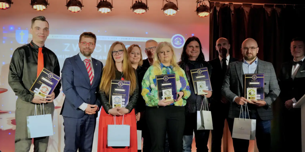 Grupowe zdjęcie dziesięciu osób na podświetlonej scenie, uchwycone podczas ceremonii wręczania nagród „Perła Mazowsza 2024”. W pierwszym rzędzie laureaci trzymają dyplomy i papierowe torby z nagrodami.