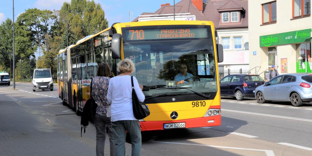 Żółto-czerwony autobus linii 710 zatrzymuje się na przystanku w centrum Konstancina-Jeziorny, gdzie na chodniku czekają pasażerowie.