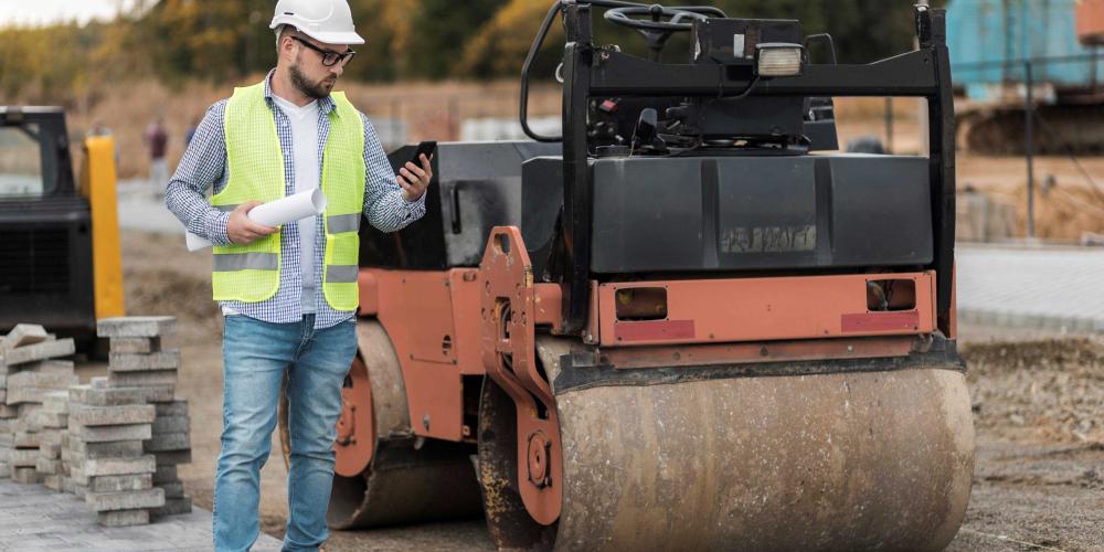Robotnik w kamizelce odblaskowej i kasku stoi obok walca drogowego na placu budowy drogi, trzymając dokumentację i telefon.