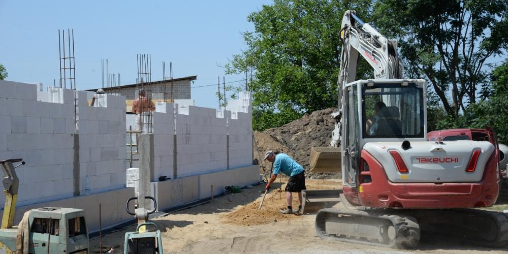Na zdjęciu trwają prace przy budowie zaplecza kompleksu sportowego. Na pierwszym planie widoczna jest koparka i pracownik przygotowujący podłoże, a w tle dwaj murarze wznoszą ściany budynku z białych bloczków. Na placu budowy znajdują się materiały budowlane oraz sprzęt wykorzystywany do realizacji inwestycji.