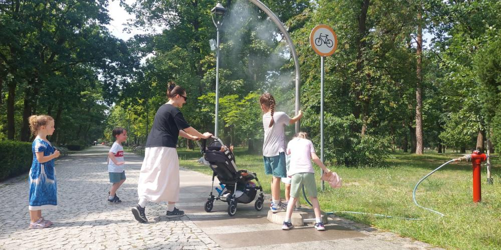 Na parkowej alei kobieta z dziećmi korzysta z bramki zraszającej, która rozpylając wodną mgiełkę przynosi ochłodę podczas upalnego dnia.