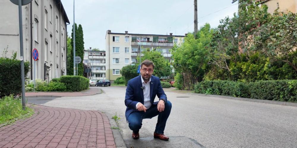 Burmistrz kuca przy wpuście kanalizacji deszczowej na osiedlowej ulicy, wskazując element infrastruktury przeznaczonej do modernizacji, a w tle widoczne są budynki mieszkalne i zieleń.