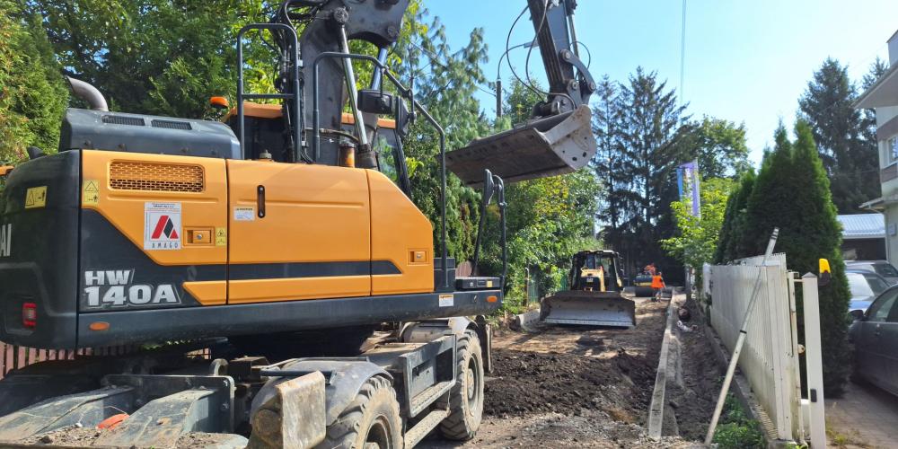 Zdjęcie przedstawia plac budowy drogi na osiedlu mieszkaniowym w słoneczny dzień. Na pierwszym planie po lewej stoi duża żółto-czarna koparka kołowa Hyundai HW140A z uniesionym ramieniem i łyżką. Widać wykop i ułożone krawężniki. W głębi, w oddali, pracuje mniejsza maszyna budowlana, a wzdłuż prawej strony ciągnie się białe ogrodzenie i rząd tui.
