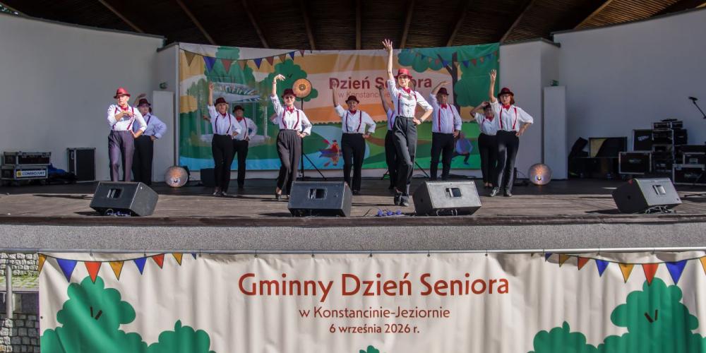 Występ artystyczny seniorów na scenie amfiteatru podczas Gminnego Dnia Seniora w Konstancinie-Jeziornie. Grupa tancerzy w białych koszulach, ciemnych spodniach, czerwonych szelkach i kapeluszach wykonuje układ taneczny z uniesionymi rękami. Z przodu sceny wisi duży baner z napisem „Gminny Dzień Seniora w Konstancinie-Jeziornie”.