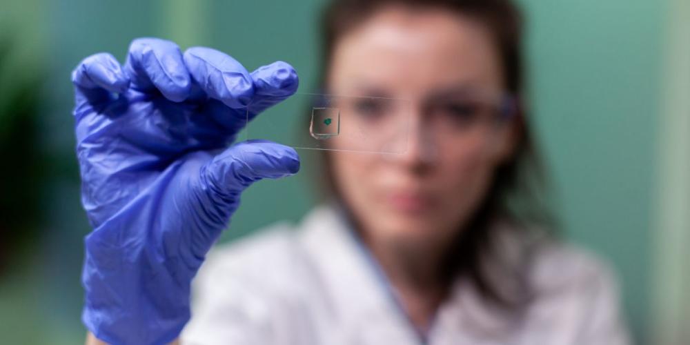 Zdjęcie przedstawia osobę w rękawiczkach laboratoryjnych analizującą próbkę na szkiełku pod mikroskopem.