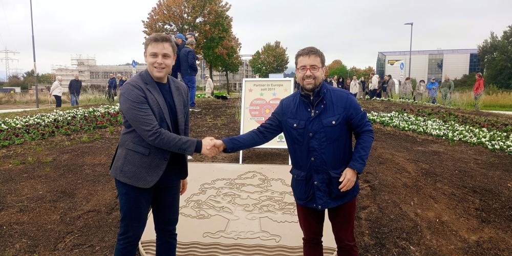 Burmistrzowie Michał Wiśniewski i Fabian Nitz ściskają dłonie na nowym rondzie w Denzlingen, przed kamiennym herbem Konstancina-Jeziorny. W tle tablica partnerska i uczestnicy uroczystości.