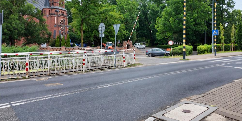 Most na ul. Mirkowskiej z zamkniętym przejazdem kolejowym, na tle zieleni i zabudowy, z widocznym oznakowaniem drogowym i przejściem dla pieszych.
