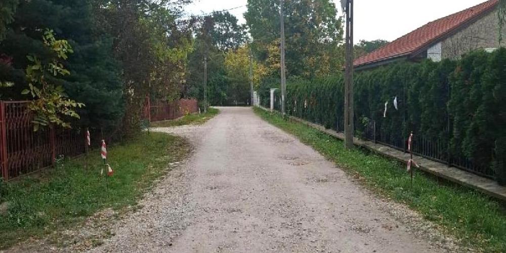 Droga gruntowa z kruszywa, biegnąca między ogrodzonymi posesjami, z drzewami i zielenią po obu stronach oraz widoczną infrastrukturą energetyczną.