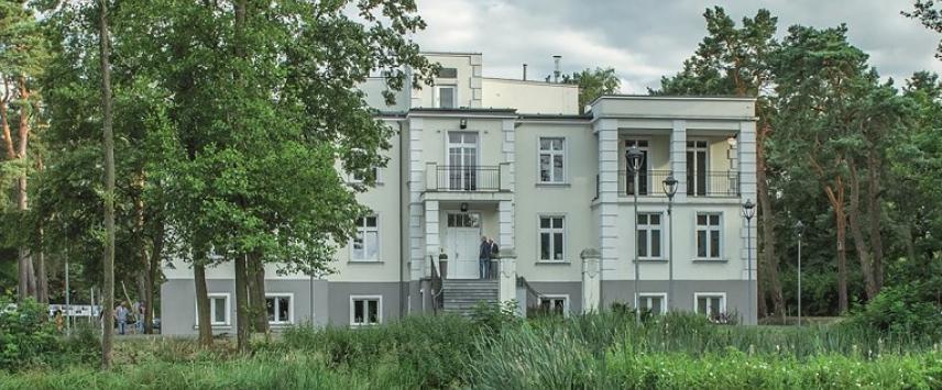 Na zdjęciu willa Hugonówka – obecna siedziba Konstancińskiego Domu Kultury (prostokątna, prosta bryła, trzy kondygnacje, dużo okien, jasna elewacje). Przed budynkiem rosną wysokie drzewa i trawy. 