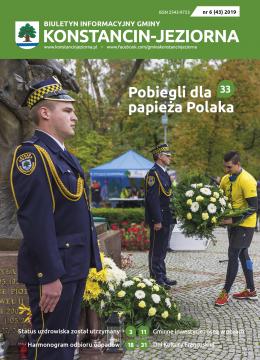 Park Zdrojowy. Dwóch mężczyzn – strażnicy miejscy, stoi przy pomniku Jana Pawła drugiego. Ubrani są w mundury Straży Miejskiej. Do pomnika podchodzi mężczyzna ubrany w sportowy strój w rękach trzyma wieniec z białymi kwiatami.