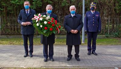 Czterej mężczyźni stojący, każdy w maseczce na twarzy, jeden trzyma wieniec biało-czerwony, jeden w mundurze straży miejskiej