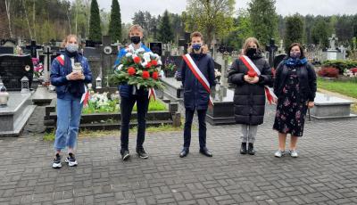 Pięc osób stojących na cmentarzu obok siebie. Dwie osoby trzymają znicz, jedna biało-czerwoną wiązankę z kwiatów . W tle las.