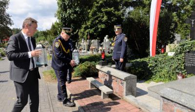 Cmentarz. Dwóch mężczyzn w średnim wieku idzie w kierunku pomnika ofiar drugiej wojny światowej. Jeden ubrany jest w garnitur, drugi ma na sobie mundur Straży Miejskiej. Każdy z nich w rękach trzyma duży znicz. Przy pomniku stoi mężczyzna ubrany w mundur Straży Miejskiej.